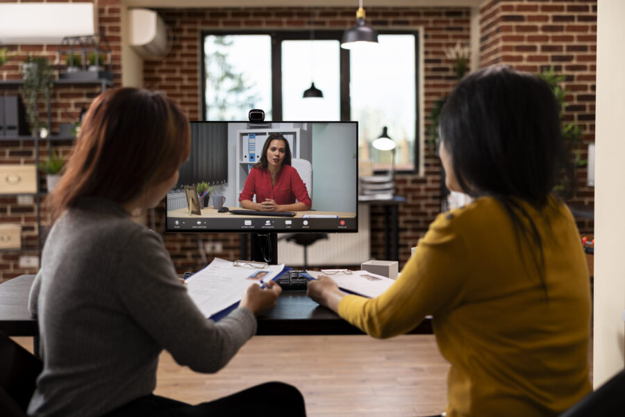 Two colleagues sitting at desk and holding clipboards, on a videoconference call with caucasian businesswoman. Female recruiters using computer for virtual meeting with manager and reviewing resumes.